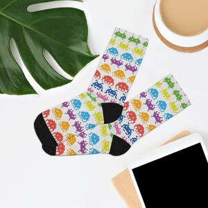May include: A pair of white socks with a rainbow pixelated alien pattern. The socks are folded and displayed on a white surface.
