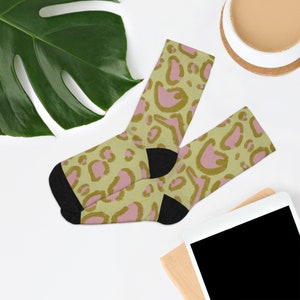 May include: A pair of crew socks with a leopard print pattern. The socks feature a light green background with pink and gold spots. The heel and toe are black. The socks are made of a soft, comfortable material.