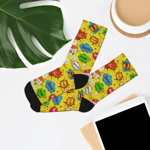 May include: A pair of yellow socks with a comic book theme. The socks feature various speech bubbles with words like "BOOM!", "POW!", and "BANG!" in red, blue, and green. The socks have black heels and toes.