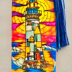 May include: A colorful stained glass bookmark featuring a lighthouse with a red door and windows, standing on a rocky shore with blue waves crashing against it. The background shows a sunset with orange, yellow, pink, and purple hues.