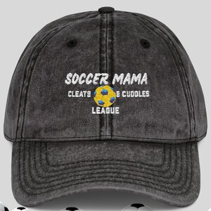 May include: A distressed black baseball cap with the text "SOCCER MAMA" in white, along with "CLEATS & CUDDLES LEAGUE". The cap features a yellow and blue soccer ball graphic. Soccer balls are at the bottom of the image.