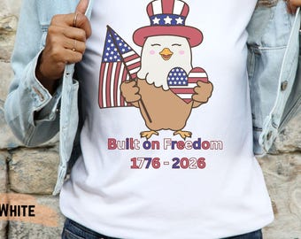 May include: White t-shirt featuring a cartoon eagle wearing a patriotic hat and holding an American flag and a heart-shaped object with the American flag design. The text "Built on Freedom 1776-2026" is printed below the eagle.