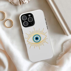 May include: White phone case featuring a gold and blue evil eye design. The case has a black camera module and is surrounded by gold rings and a beige scrunchie. The evil eye is a symbol of protection.