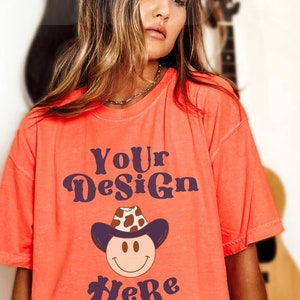 May include: A bright orange t-shirt with the text "Your Design Here" in purple. The design features a cartoon smiley face wearing a brown and white cowboy hat.