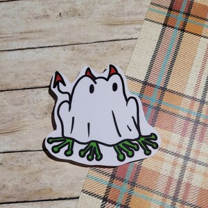 May include: White ghost frog sticker with red devil horns and green webbed feet.