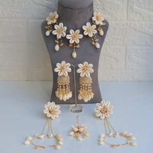 Op de afbeelding: Een witte schelpbloemen ketting en oorbellen set. De ketting heeft een rij witte schelpbloemen met gouden accenten. De oorbellen zijn hangende oorbellen met een witte schelpbloem en gouden accenten. De oorbellen hebben een lange ketting met witte schelp kralen.