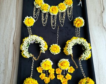May include: A collection of yellow floral jewellery, including a necklace, earrings, bracelets, and a headpiece, displayed on a black surface. The set features yellow flowers and white accents, with gold chains and details.