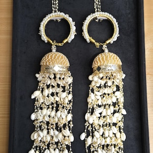 May include: A pair of ornate, dangling earrings. Each earring features a floral top, a pearl-lined hoop, and a domed, beaded base. White seashells cascade from the base, creating a layered, textured effect. The earrings are displayed on a black surface.