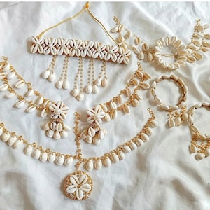 Könnte beinhalten: Ein Set aus weißem Muschel-Schmuck mit goldenen Akzenten. Das Set beinhaltet eine Halskette, ein Choker, Ohrringe, ein Stirnband und ein Armband. Der Schmuck ist aus weißen Muscheln und einer goldenen Kette gefertigt.