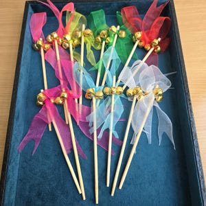 May include: A collection of colorful decorative bell wands. Each wand features a small gold bell, a sheer ribbon bow, and a wooden stick. The ribbons are in shades of pink, red, green, blue, and white. The wands are arranged in a dark blue tray.
