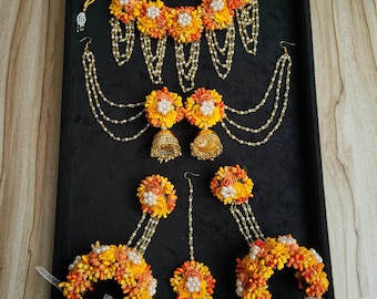 May include: A full floral jewellery set in yellow, orange, and white. The set includes a necklace, earrings, and bracelets. The jewellery features delicate flower arrangements with pearl accents and gold-coloured chains, displayed on a black surface.