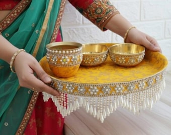 May include: A yellow decorative tray with gold bowls and a cup, adorned with pearls and floral accents. The tray has a beaded fringe. The items are held by a person wearing a red and green outfit.