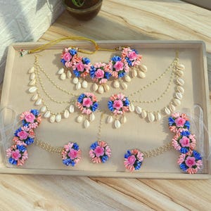 Könnte beinhalten: Ein floraler Schmuck-Set mit Halskette, Ohrringen und Armbändern. Das Set umfasst rosa, blaue und pfirsichfarbene Blumen, weiße Muscheln und perlenartige Perlen. Der Schmuck ist auf einem beigen Tablett angeordnet.