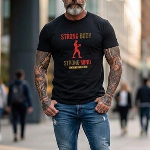 Può includere: T-shirt nero con la scritta "STRONG BODY STRONG MIND" in rosso e oro. La maglietta presenta una grafica di una persona in posa da pugilato. La persona indossa jeans blu con ginocchia strappate.