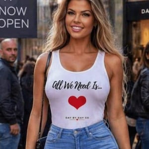 May include: A woman wearing a white tank top with the text "All We Need Is..." and a red heart graphic. She is also wearing blue jeans. The sign behind her says "NOW OPEN".