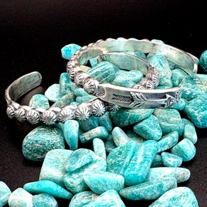May include: Two silver-toned cuff bracelets with detailed designs. One features a row of round, textured embellishments, while the other showcases an arrow and feather motif. The bracelets rest on a bed of turquoise-colored stones against a black background.