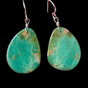 May include: A pair of turquoise teardrop earrings with silver-colored hooks. The earrings have a mottled appearance with shades of green and brown, set against a black background.