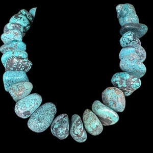 May include: A necklace composed of turquoise-colored, irregularly shaped stones. The stones have a mottled appearance with shades of blue, green, and gray. The necklace is set against a black background, highlighting the vibrant colors of the stones.