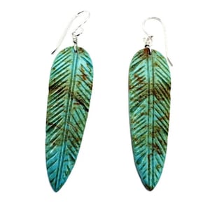 May include: A pair of dangle earrings featuring carved turquoise-colored stone pendants shaped like elongated leaves. The pendants display a textured, veined surface with brown inclusions and are suspended from simple silver-toned metal ear wires.