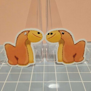 May include: Two orange dinosaur stickers with yellow heads and smiling faces. The stickers are placed on a light blue tiled surface, with a pink background. The stickers are facing each other.