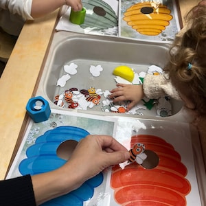 May include: A children's activity set with a beehive theme. The set includes a blue and orange beehive with a hole in the center, a green beehive, a yellow beehive, and a variety of bee cutouts. The bee cutouts are scattered around a white sink.