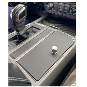 May include: Interior car console with a black surface and a silver trim. A silver and black gear shift is visible, along with a small, silver, cylindrical object on the console. The console is part of a vehicle's dashboard.
