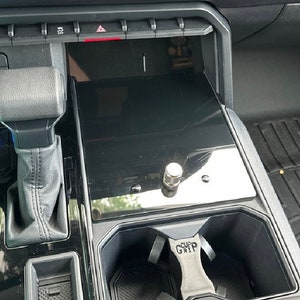 May include: A black car interior with a gear shifter, a cup holder, and a black surface with a silver knob. The cup holder has a black cup grip with the text "CUP GRIP" on it.