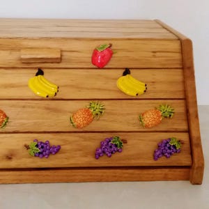 May include: A rectangular wooden bread box with a slanted front lid featuring a central handle. The front panel is decorated with rows of colorful, raised fruit appliques, including red apples, yellow bananas, pineapples, and purple grapes.