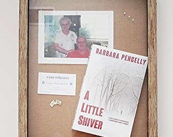 May include: A rustic wooden shadow box frame containing a photograph of two people, a business card for Barb Pengelly, two small gold-toned flag pins, and a book titled A Little Shiver by Barbara Pengelly with a winter forest cover design.