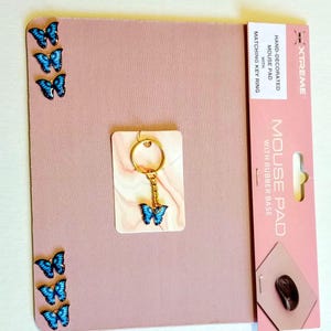 May include: A pink mouse pad with a matching key ring. The mouse pad has a rubber base and is decorated with blue butterfly accents. The key ring features a gold chain and a blue butterfly charm. The packaging reads "HAND-DECORATED MOUSE PAD WITH MATCHING KEY RING".