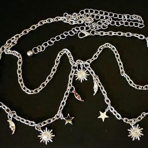 May include: Silver-toned body chain with celestial charms. The chain features sun, moon, and star pendants, along with feather charms. The chain is displayed on a black background.
