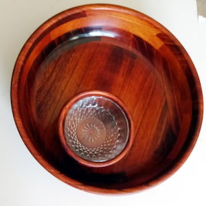 May include: A wooden salad bowl with a glass insert. The large bowl is made of dark wood with a reddish-brown hue, and the smaller insert is clear glass with a starburst design. The bowl is round and has a smooth finish.