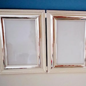 May include: A silver-colored, hinged double picture frame. The frame has a textured border and a bright white interior. The frame is displayed against a blue background.