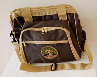 May include: A dark brown rectangular bag with beige trim, two top handles, and an adjustable shoulder strap. The bag features two zippered front compartments and a circular embroidered patch depicting a green tree with brown roots on the front.