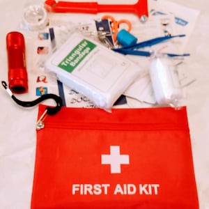 May include: A red zippered pouch with a white cross and the text "FIRST AID KIT". The pouch is open and contains a variety of first aid supplies, including a triangular bandage, gauze, a flashlight, and a safety hammer.