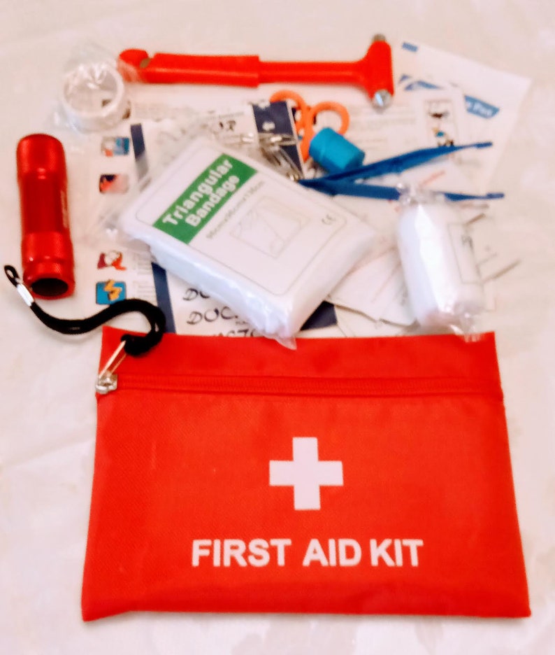 May include: A red zippered pouch with a white cross and the text "FIRST AID KIT". The pouch is open and contains a variety of first aid supplies, including a triangular bandage, gauze, a flashlight, and a safety hammer.