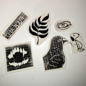 Könnte beinhalten: Eine Sammlung von sechs schwarz-weißen Linoldrucken auf cremefarbenem Papier. Die Motive zeigen ein Farnblatt, ein menschliches Auge, einen Vogel, einen Cartoon-Hund, ein Gebiss mit spitzen Zähnen und ein Rechteck mit dem Text FUCKED.
