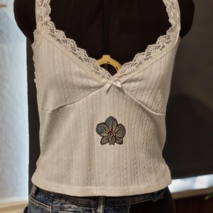 Könnte beinhalten: Ein weißes, geripptes Camisole mit V-Ausschnitt, zarter weißer Spitzenborte und einer kleinen Schleife in der Mitte. Das Kleidungsstück zeigt mittig einen aufgestickten blauen Orchideen-Aufnäher. Der Stoff hat eine vertikale Fischgrätstruktur.