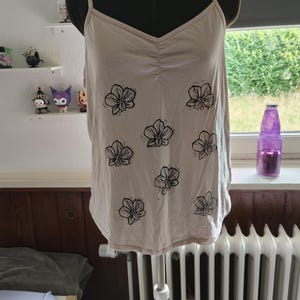 Könnte beinhalten: Ein beiges Camisole mit V-Ausschnitt, dünnen Spaghettiträgern und einem gerafften Detail in der Mitte. Das Kleidungsstück ist mit einem sich wiederholenden Muster aus schwarzen, tuscheartigen Blumenillustrationen auf der Vorderseite bedruckt.