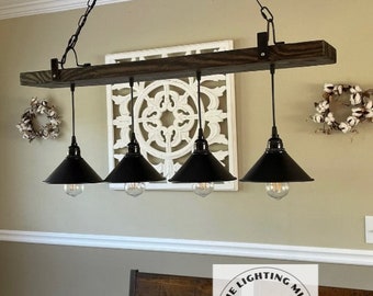 May include: A rustic farmhouse style chandelier with four black metal shades and a wooden beam. The chandelier is hanging from a chain with a pulley wheel. The chandelier is hanging over a dining table with a white tablecloth and a green runner. The table is set with plates, glasses, and pumpkins. The text "The Lighting Mill Handcrafted Goods" is visible in the lower right corner of the image.