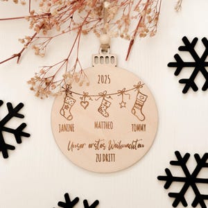 Runde Weihnachtskugel aus hellem Holz mit Aufhängung und sandfarbenem veganem Lederband samt Holzperle. Auf der Vorderseite sind oben „2025“, darunter drei hängende Strümpfe und die gravierten Namen „JANINE“, „MATTHEO“, „TOMMY“ sowie der Schriftzug „Unser erstes Weihnachten zu dritt“ zu sehen; dekoriert auf weißer Holzfläche mit schwarzen Schneeflocken-Elementen und zarten getrockneten Blüten.