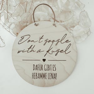 Könnte beinhalten: Runder, hellfarbiger Holzschild mit brauner Lederschlaufe zum Aufhängen. Das Schild trägt den Text "Don't google with a Kugel" über einem Herz und den Text "DAFÜR GIBT ES HEBAMME LENA!" in braunen Buchstaben.