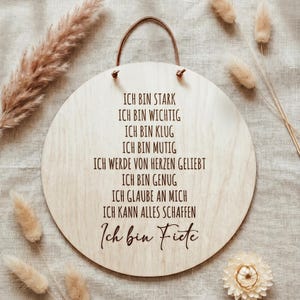 Könnte beinhalten: Runder Holzschild mit Lederaufhängung. Der Text "ICH BIN STARK, ICH BIN WICHTIG, ICH BIN KLUG, ICH BIN MUTIG, ICH WERDE VON HERZEN GELIEBT, ICH BIN GENUG, ICH GLAUBE AN MICH, ICH KANN ALLES SCHAFFEN, Ich bin Fiete" ist in dunkler Schrift geschrieben.