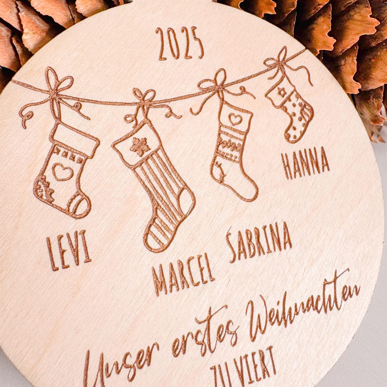 Puede incluir: Adorno navide&ntilde;o redondo de madera con grabados. Muestra el a&ntilde;o "2025" y cuatro calcetines colgantes, cada uno con un nombre. El texto "Unser erstes Weihnachten ZU VIERT" est&aacute; grabado en la parte inferior, creando un ambiente festivo.