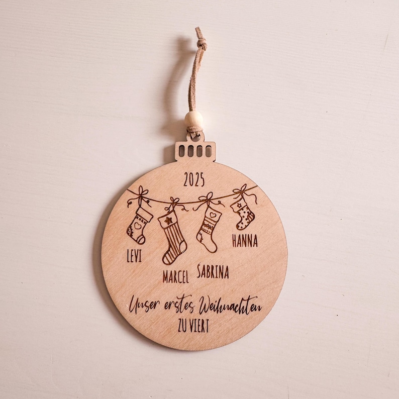 Puede incluir: Adorno navide&ntilde;o redondo de madera con una cuerda para colgar. El adorno presenta cuatro calcetines colgados de una cuerda, con nombres y el a&ntilde;o 2025. Tambi&eacute;n se incluye el texto "Unser erstes Weihnachten zu Viert".