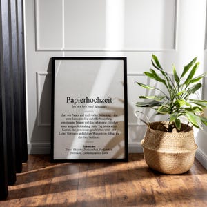 May include: A framed print with the German word "Papierhochzeit" (paper wedding) in black text. The print is in a black frame and is displayed on a wooden floor. A woven basket with a green plant sits to the right.