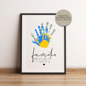Può includere: Stampa artistica incorniciata con impronte di mani colorate in blu, giallo e verde sovrapposte. La parola "familie" è scritta in corsivo sotto, con testo aggiuntivo e un simbolo a forma di cuore. È presente anche un'opzione di download digitale.