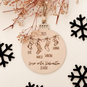 Runde Weihnachtskugel aus hellem Holz mit Gravur „2025“, darunter vier hängende Strümpfe mit den Namen „LEVI“, „MARCEL“, „SABRINA“ und „HANNA“. Unten steht der Schriftzug „Unser erstes Weihnachten zu viert“. Oben Aufhängung mit sandfarbenem veganem Lederband und Holzperle. Der Anhänger liegt auf heller Holzfläche, dekoriert mit schwarzen Schneeflocken-Elementen und zarten getrockneten Blüten – festliche Familien-Deko im Boho-Stil.