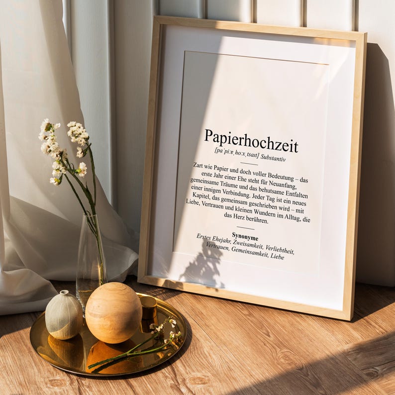 May include: A framed print with the German word "Papierhochzeit" (paper wedding) in a minimalist style. The print is in a light wood frame, and the text is in black. A gold tray with decorative spheres and a vase of flowers is in the foreground.