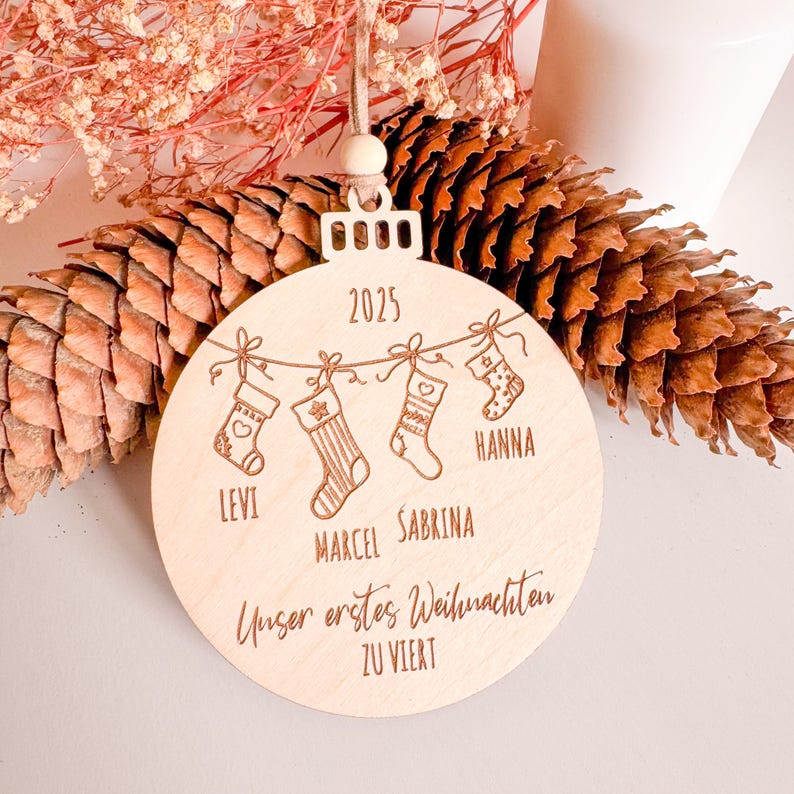 Runde Holz-Weihnachtskugel mit Gravur „Unser erstes Weihnachten zu viert“, Jahr 2025 und vier personalisierten Namen. Motiv mit aufgehängten Strümpfen, Herz und Stern. Christbaumanhänger mit veganem Lederband und Holzperle, dekorativ präsentiert mit Tannenzapfen und getrockneten Blüten.